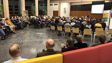 Mathildezaal gemeentehuis volle zaal met mensen op banken en stoelen die luisteren naar spreker op podium met flipover