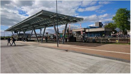 groot zonnepanelendak op een busstation waaronder mensen wachten. Op de achtergrond het stationsgebouw