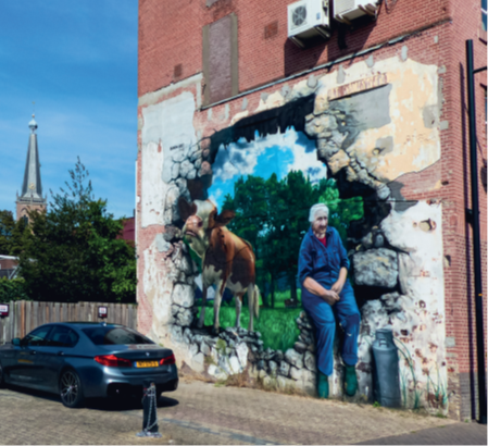 Muurschildering van boerin Anna zittend naast een melkbus. Links van haar een koe. Op de achtergrond de kerktoren. Op de voorgrond een geparkeerde auto.